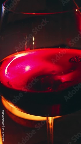 Vertical video. Hand Holding Glass of Red Wine Beside Candle with Deep Red Light Reflection on Surface, Cozy Night Relaxation Concept