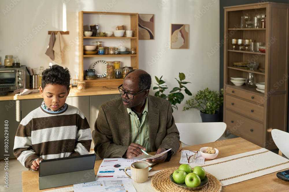 Fototapeta premium Boy using tablet at kitchen table while senior Black man sitting beside him holding documents and pen, both focused on paperwork and technology in home setting
