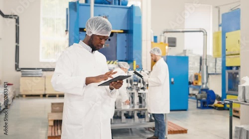 Handsome African American male worker in protective coat using tablet device and scrolling with finger. Using gadget. Focused man monitoring production process at modern manufacturing factory.