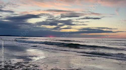 Powerful clouds and vivid sunset colors reflect over rolling ocean waves, creating a dynamic and dramatic seaside scene.