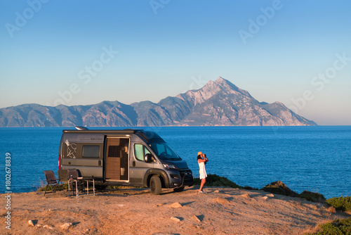Woman enjoys the Aegean view beside a camper van with Mount Athos in the background. Ideal for: vanlife travel, outdoor brands, road-trip blogs, Greece tourism. Colours: blue, beige, silver, gold.