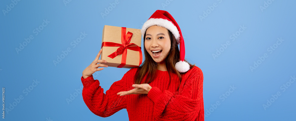 Obraz premium A young woman wears a bright red sweater and Santa hat while joyfully holding a wrapped present. She smiles widely, sharing holiday cheer in a cheerful atmosphere.