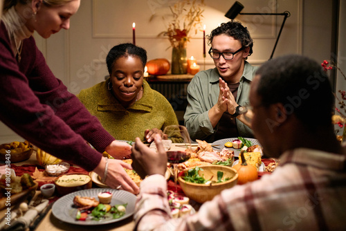 Group of diverse young adults and Black woman sitting around table sharing Thanksgiving dinner, eating and talking, hands reaching for food, festive meal setting, autumn decorations visible