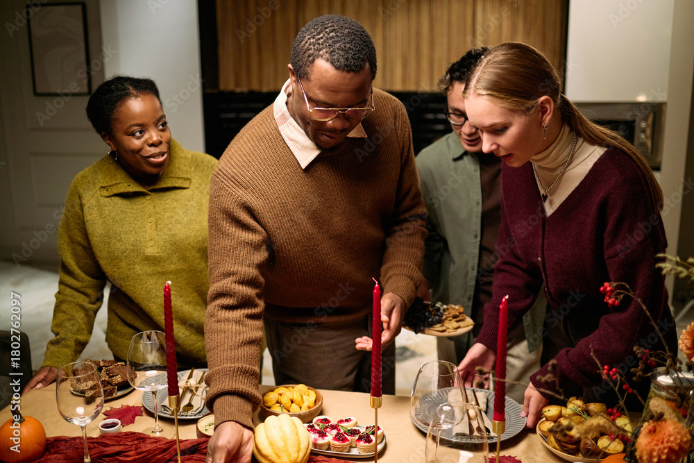 Obraz premium Group of young adult multiethnic friends gathering around table serving traditional Thanksgiving dinner food, Black man reaching for dish while Caucasian woman and others smiling and interacting