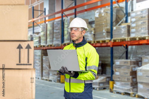 Warehouse worker wearing PPE taking inventory using a laptop in an industrial warehouse.