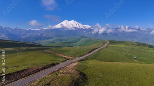 The car is driving along a mountain road in the Caucasus, towards the foot of Mount Elbrus in the Dzhily-Su tract, with the snow-capped peak of Mount Elbrus on the horizon