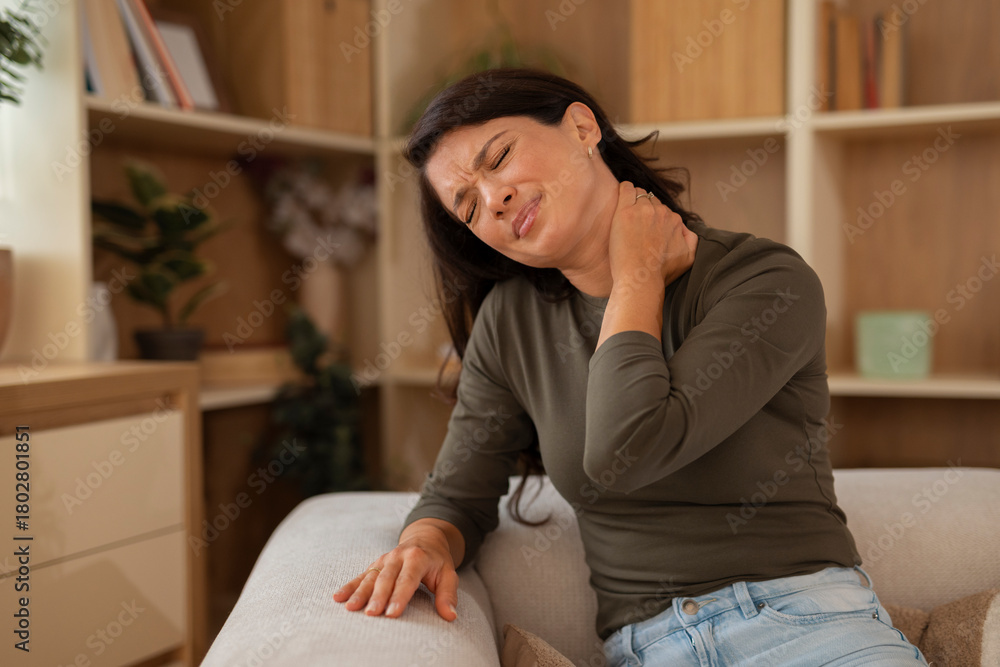 Fototapeta premium Young woman touching her neck with a painful expression, showing neck pain while sitting at home. 