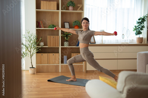 A woman exercising at home, performing a lunge with extended arms and dumbbells on a yoga mat in a bright living room.  
