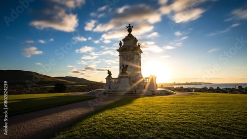 Time-lapse of the sun setting behind a monument on a grassy hill overlooking a city and the sea