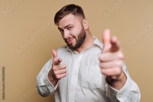 portrait of young confident man in a white shirt playfully points fingers towards the camera on beige background