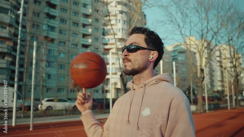 A young brunette Caucasian man wearing sunglasses and headphones confidently spins a basketball on his finger while on a striking red outdoor court with communistic buildings in the background.