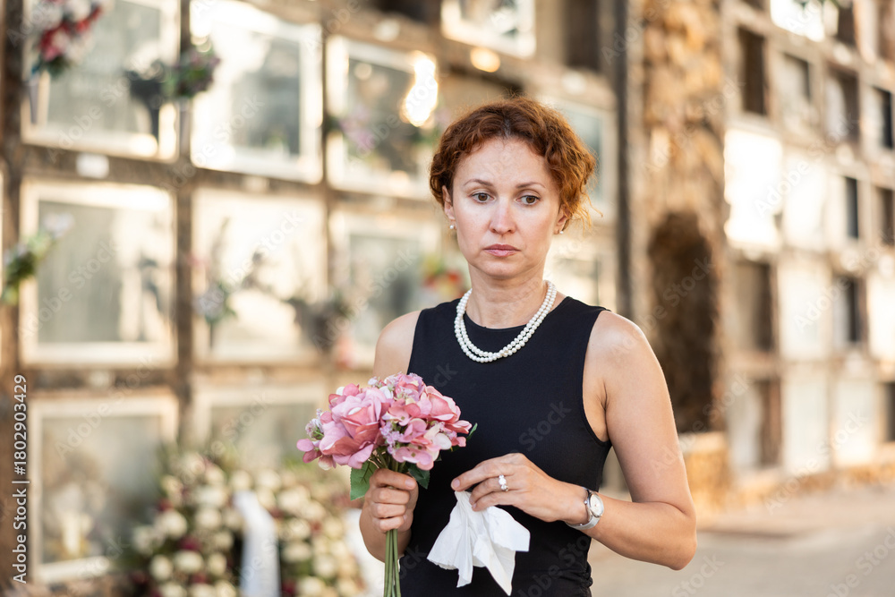 Fototapeta premium Sorrowful middle aged woman in black dress standing in cemetery holding bouquet of flowers with reflective and somber expression against backdrop of stone memorial walls