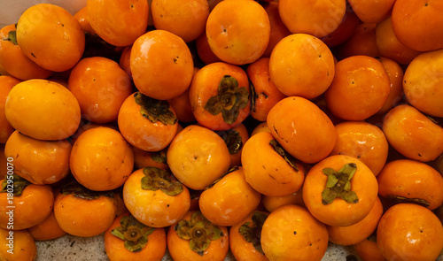 Fotografi Freshly harvested ripe orange persimmons piled up at a market stall, showing their smooth skin and calyx