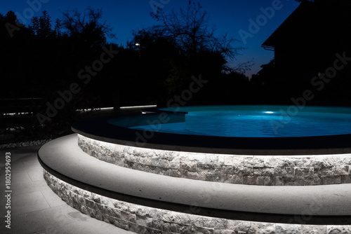 A modern outdoor pool glowing at night with vibrant LED lighting inside and around the water, creating a luxurious, serene, and atmospheric scene ideal for relaxation and nightlife visuals.