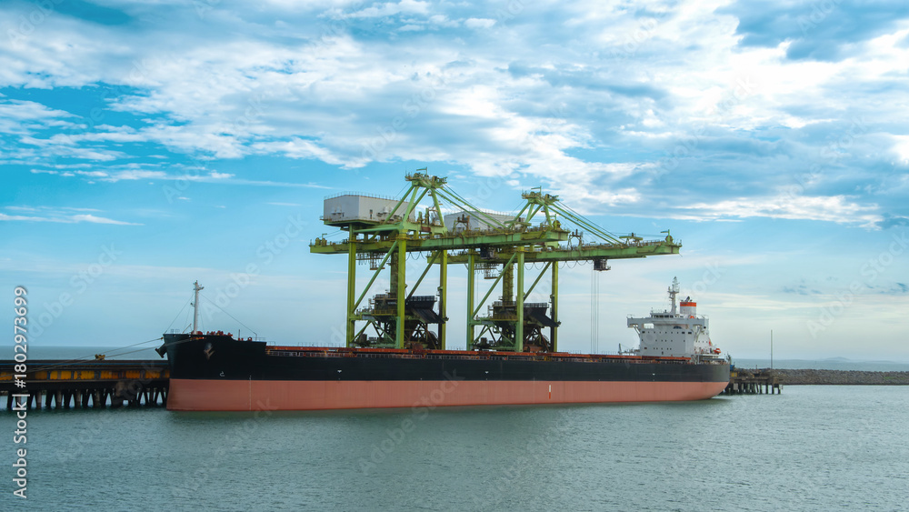 Fototapeta premium Large cargo ship docked at a pier beneath massive green port cranes set against a blue cloudy sky. Transportation and logistics