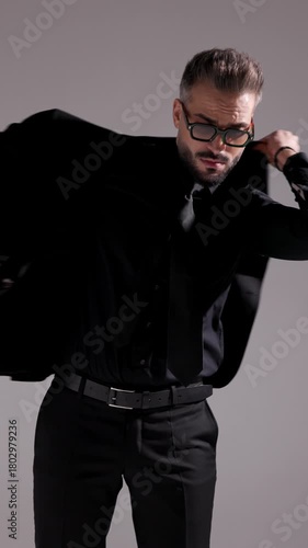 Closeup of a cool man in sunglasses and all black outfit putting on his jacket and smiling on grey background.