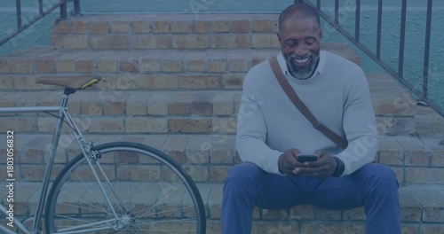 Sitting cyclist in gray sweater checking phone on brick steps, with bicycle, satchel, copy space
