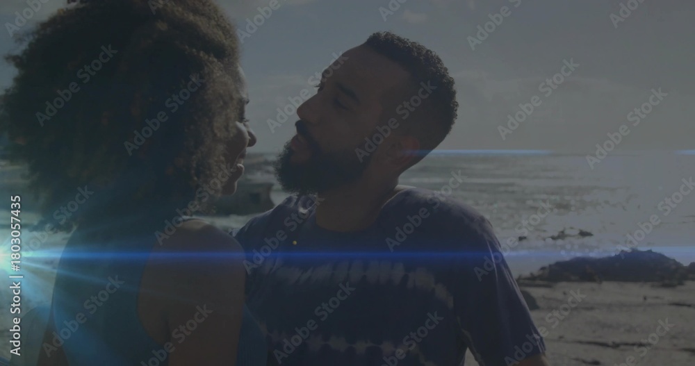 Naklejka premium Embracing couple leaning at rocky shoreline with ocean horizon, sun flare, tank top and tie-dye tee