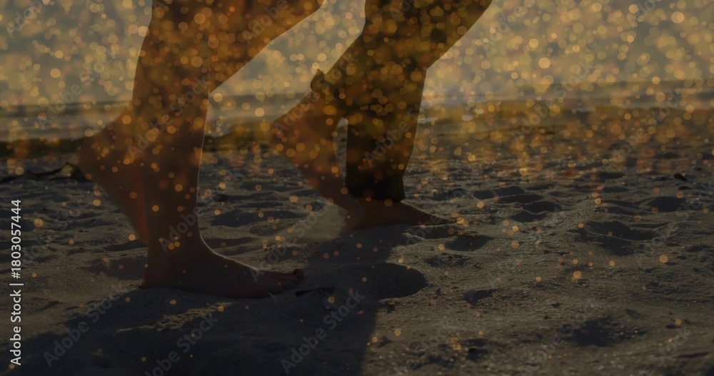 Fototapeta premium Walking two adults' bare feet and legs leaving footprints along sandy shore at sunset, golden bokeh