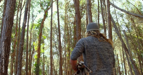 Standing man holding bundle of sticks, wearing knit beanie and sweater in pine forest