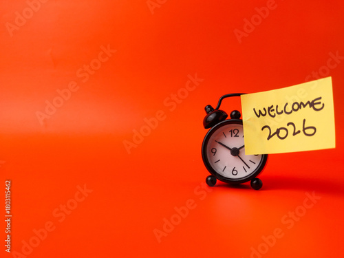 Black alarm clock and sticker note with the word WELCOME 2026 on a orange background