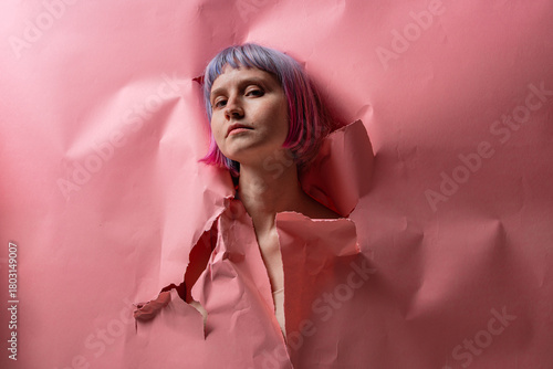 A woman with vibrant pink hair emerges from a torn pink backdrop in a bright art studio. Her confident posture showcases a blend of strength and creativity, symbolizing breaking free from constraints