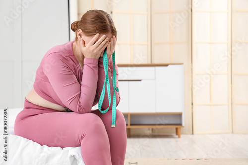Upset sporty woman with excess weight and tape measure sitting in bedroom