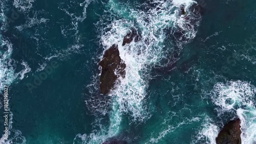 Bright blue waves surge and crash against dark jagged rocks, creating a beautiful contrast in this lively seascape filled with energy and nature's raw beauty.