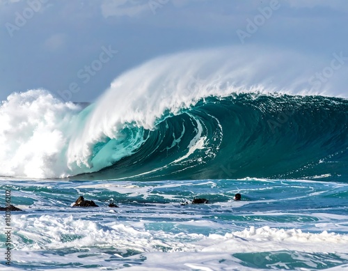Fototapeta Naklejka Na Ścianę i Meble -  A majestic ocean wave crests over rocks, showing a powerful turquoise interior and white foam. The sky is partially cloudy