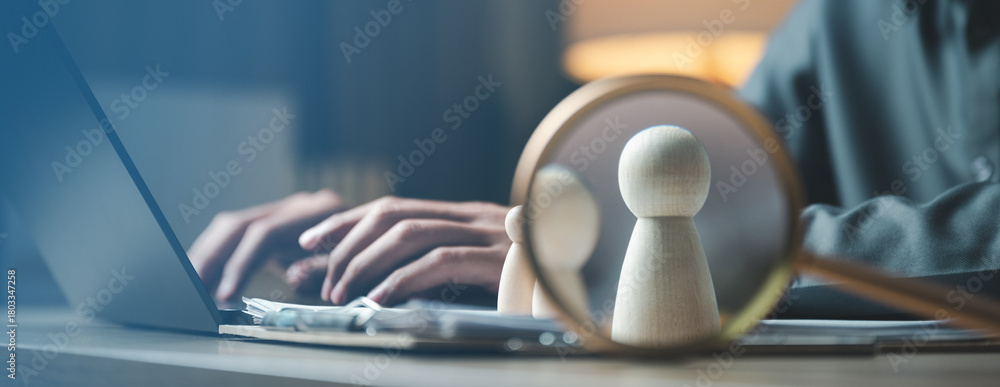 Fototapeta premium Magnifying glass focusing on wooden figures with a person typing on a laptop, representing recruitment, talent search, HR evaluation, and candidate selection processes.