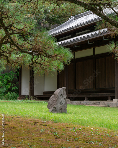 A tranquil scene featuring a traditional wooden guardhouse and a moss-covered stone marker with kanji calligraphy, framed by the dense boughs of a Japanese pine tree
