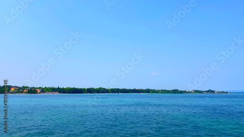 Peaceful view at Lago di Garda from a ferry cruising the lake's fluttering wavy blue surface, buildings, trees and thicket on the distant shores in the background, outline of mountains, a seamless sky