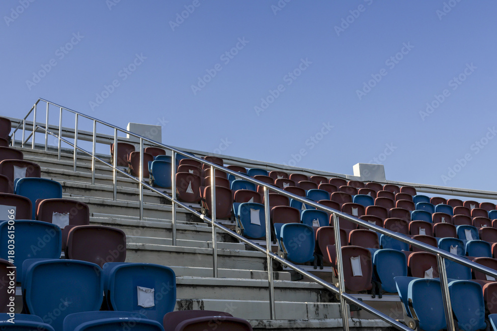 Obraz premium Empty blue seat in row at an outdoor stadium under clear sky. quiet and solitary scene showing grandstand, aisle, and metal railing for perfect sport background