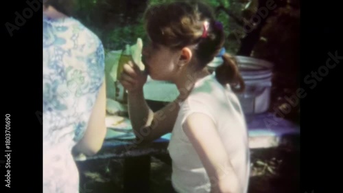Teenage girl eating snacks. Nutrition in teenage years. Young girl with ponytail enjoying sandwich at outdoor picnic table with family during summer. Old film. Archival retro 1960s. Vintage archive.