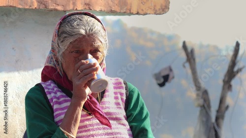 Old Indian rural woman around 80 to 85 years enjoying a warm cup of tea outdoors at her village home, sitting peacefully in a natural mountain setting.