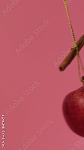 A red apple swings from side to side like a pendulum on a shiny gift rope with a cinnamon stick on a pink background. A beautiful display of flavors.