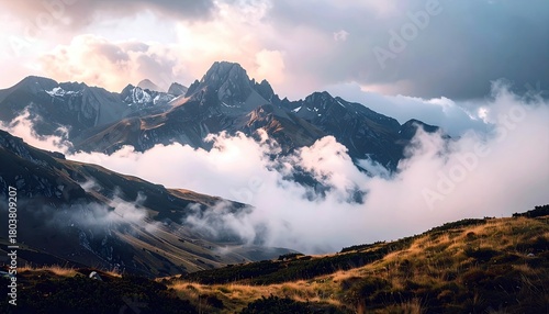 A dramatic landscape of rugged mountain peaks emerging from swirling clouds, with sunlit golden grasses in the foreground.