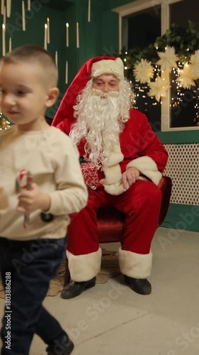 Santa Claus sits with two children holding Christmas gifts in a festive room. Vertical video.
