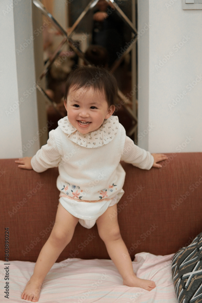 Fototapeta premium Happy Asian toddler girl playing and standing on a sofa at home, laughing and having fun.