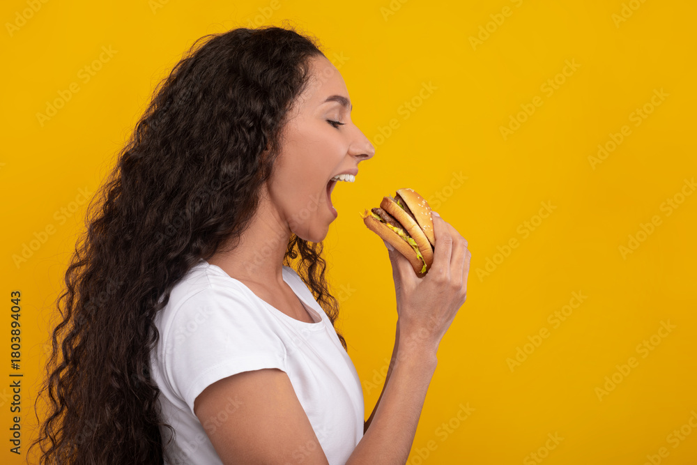 Naklejka premium Smiling female model bites into a tasty burger with excitement. She is in a casual outfit against a vibrant yellow wall. Her joyful expression shows her love for this delicious fast food snack.