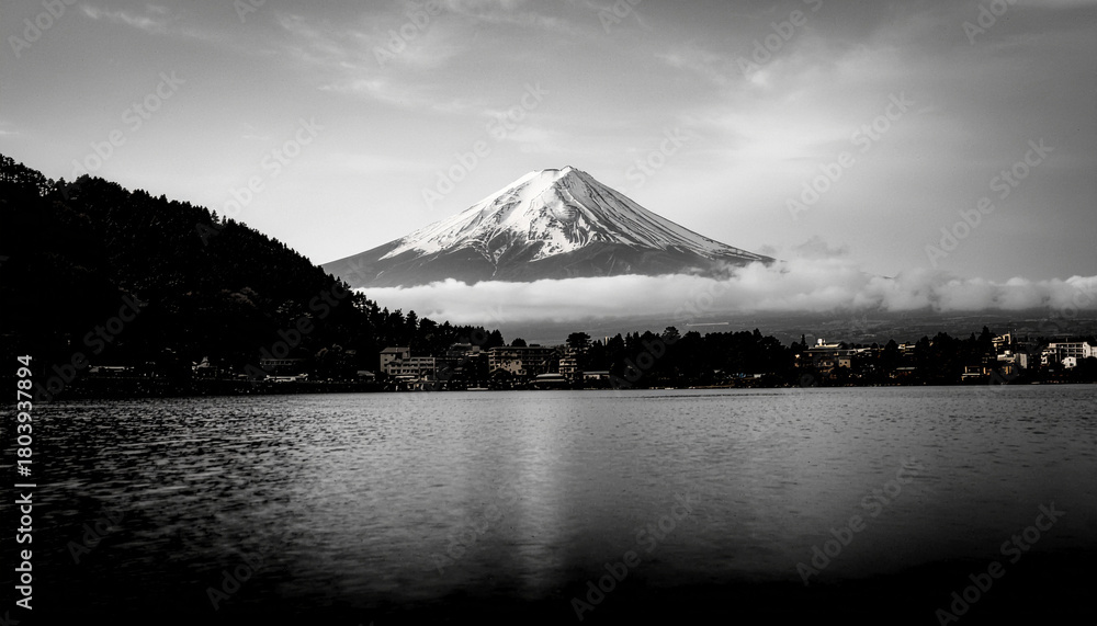Fototapeta premium A dramatic black and white landscape of snow-capped Mount Fuji, partially shrouded in mist, reflected over a calm lake near a distant town.