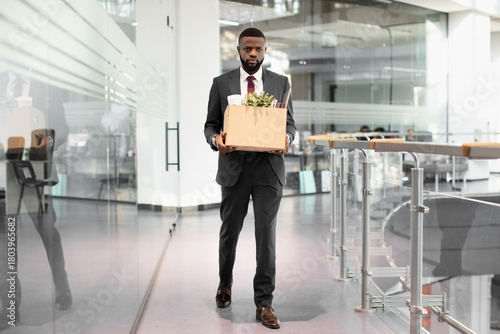 Unhappy young black man in suit employee got fired from job, walking by modern office, holding box with his belongings, looking angry and upset, copy space, full length shot. Unemployment concept
