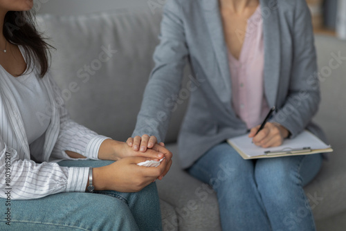 A female psychotherapist offers emotional support to a young woman by holding her hand during a therapy session in a clinic. The setting appears calm and inviting, focused on healing.