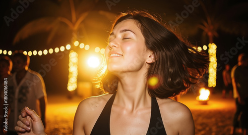 Young woman with her eyes closed, smiling while dancing outdoors at night under warm, golden lighting from string lights and a bonfire.