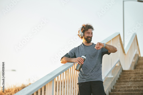 Canvastavla A man with headphones stands on a staircase by the river, checking his fitness watch while holding a water bottle