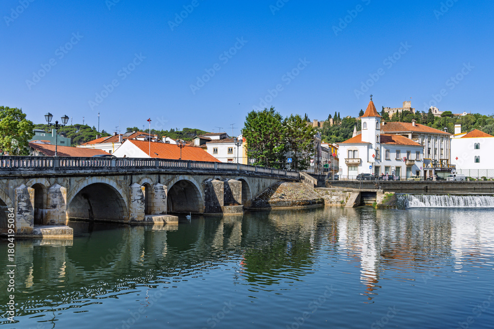 Obraz premium A scenic view of the River Nabao in Tomar, Portugal, featuring a historic arched stone bridge and the city's cityscape under a clear blue sky.