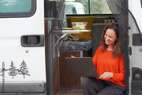 Teleworking and travel among middle-aged women. Smiling woman working on her laptop at the door of her camper van.