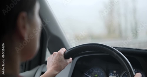 Over the shoulder view of a woman driving a car during heavy rain. Hands on steering wheel, windshield wipers clearing water from the glass. Bad weather driving.