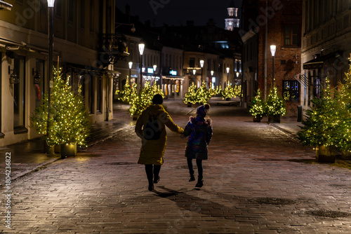 Mother and daughter walking through Kaunas old town Christmas lights at night, festive trees along cobbled street