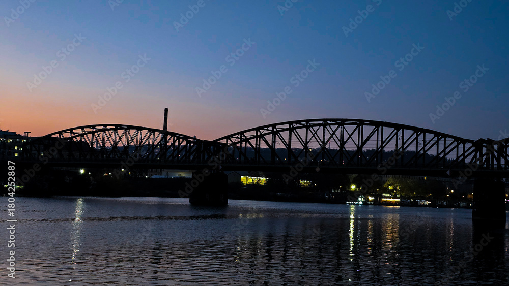 Naklejka premium The silhouette of a metal city bridge against the evening sky during blue hour. Reflections of lights on the river surface.
